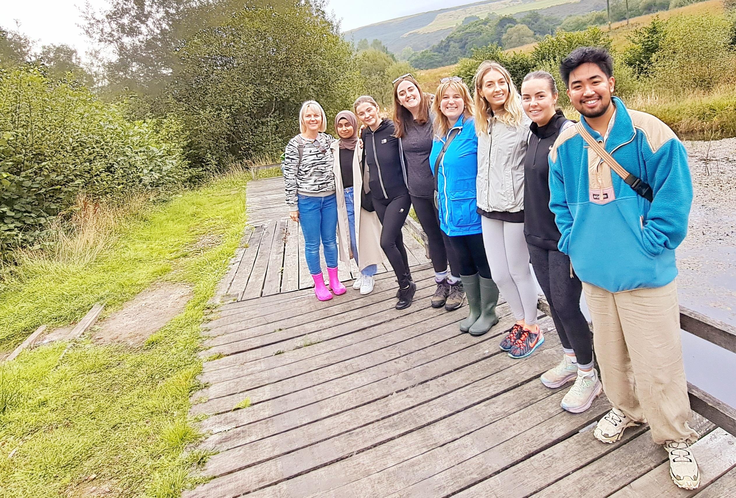 A photo of the SVC team. A group of seven people standing on a wooden path in a scenic outdoor setting with trees and hills in the background.